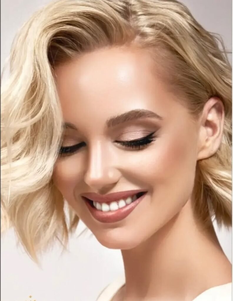 Close-up of a woman with flawless natural makeup, softly styled blonde hair, defined brows, and neutral-toned eye makeup in a clean, luxury beauty setting.